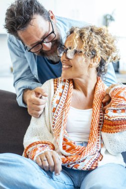 Olgun bir çift, rahat ve neşeli bir yüz ifadesiyle birbirlerine gülümsüyor, güzel döşenmiş bir oturma odasındaki rahat bir koltukta rahatça oturuyor, pozitif ve sevgi dolu bir ilişkiyi somutlaştırıyor.