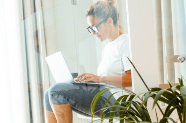 A woman in casual clothing uses a laptop while sitting on an internal staircase in a modern home, managing her online business and working from home in a creative and comfortable setting clipart