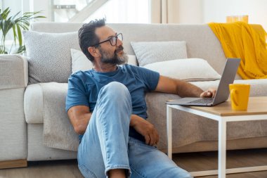 50 yaşında bir adam oturma odasında yerde oturuyor, günlük kıyafetler giyiyor, internette gezinmek için küçük bir masaya dizüstü bilgisayar koyuyor, sosyal medyayla meşgul oluyor ve boş zamanlarında internette çalışıyor.