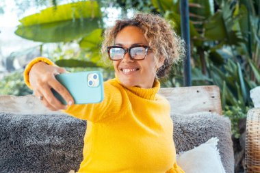 Güneşli bir günde arka bahçesinde oturan kıvırcık saçlı, sarı bir kazak ve kot giyen akıllı telefonuna gülümseyen ve video aracılığıyla arkadaşlarıyla sohbet eden şık bir kadın.