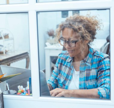 Ev ofis ortamında dizüstü bilgisayarı üzerinde çalışan orta yaşlı bir kadın, pencereden bakılıyor, çevrimiçi sosyal medya veya mesleki iş görevleriyle uğraşıyor, daktilo ediyor, tarıyor, içeriği düzenliyor ve yönetiyor