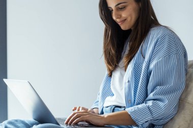 Genç, modern, yetişkin bir kadın kanepeye oturmuş dizüstü bilgisayarını ve internet bağlantısını iş ve eğlence için kullanıyor. Uzaktan kumandayla çalışan insanlar yeni faaliyet konsepti yaşam tarzı. Defterde mutlu kadın yazıları