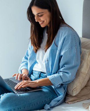 Laptop 'a yazı yazan genç bir kadının yan görüntüsü. Evde, yatakta rahat bir şekilde oturuyor. Uzaktan çalışan ya da öğrenci yaşam tarzı. İnsanlar ve internet teknolojisi. Gündelik kıyafet, modern kadın.