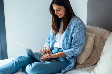 Laptop 'a yazı yazan genç bir kadının yan görüntüsü. Evde, yatakta rahat bir şekilde oturuyor. Uzaktan çalışan ya da öğrenci yaşam tarzı. İnsanlar ve internet teknolojisi. Gündelik kıyafet, modern kadın.