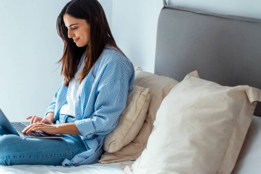 Şirin, modern bir kadın evinde rahat bir şekilde yatağında oturuyor. İnternetteki yeni iş kurma konsepti için dizüstü bilgisayar ve kablosuz internet bağlantısı kullanıyor. İnsanlar sohbet ediyor ve boş zamanların tadını çıkarıyor.