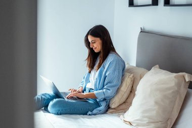 Şirin, modern bir kadın evinde rahat bir şekilde yatağında oturuyor. İnternetteki yeni iş kurma konsepti için dizüstü bilgisayar ve kablosuz internet bağlantısı kullanıyor. İnsanlar sohbet ediyor ve boş zamanların tadını çıkarıyor.