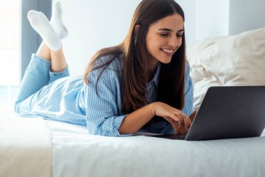 Evde ya da otel odasında dizüstü bilgisayar kullanarak yatıp ekranda gülümseyen neşeli bir genç kadın. İnternette arkadaşlarla sohbet etmek ya da video bağlantısı kurmak. Boş zaman aktivitesi kavramı