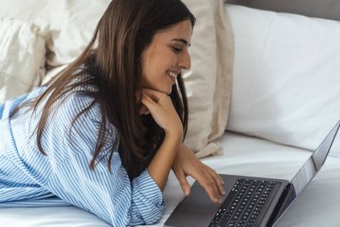 Rahat bir yatakta uzanan ve dizüstü bilgisayarla çalışan gündelik giysili çekici genç bir kadın. Mutlu bir kadın bilgisayarla eğleniyor, genç bir kadın internette geziniyor, internetten iş fırsatları arıyor, oyunlar oynuyor.