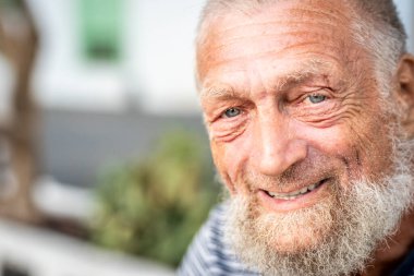 Yaşlı adam dışarıda dinleniyor, mavi gözlü ve beyaz saçlı gülümsüyor, emekliliğin ve doğanın tadını çıkarıyor, sakin ve neşeli bir ifadeyle yaşlanmanın güzelliğini temsil ediyor, ve sağlık ve sağlığa odaklanmış bir sakalı var.