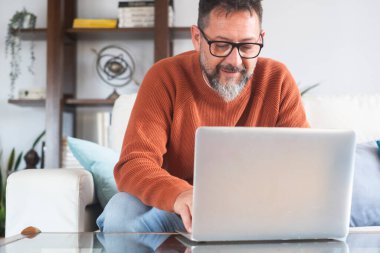 Mutlu, orta yaşlı, gözlüklü, bilgisayar notları kullanarak dizüstü bilgisayara bakan ve evde oturup Webinar izleyen olgun bir adam. Melez işi, elkazanımcılık, çevrimiçi öğrenme kavramı.