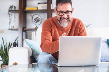 Orta yaşlı bir adam gözlük takıyor ve rahat bir şekilde evdeki bir kanepede oturuyor. Uzaktan kumandalı bir dizüstü bilgisayar üzerinde çalışıyor, serbest işleri kişisel online eğlence aktiviteleriyle birleştiriyor.