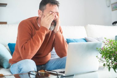 Yorgun yetişkin bir adam gözlük tutarken gözlük tutarken acı hisseder bilgisayar işinden yorgun düşer, stresli bir adam baş ağrısı çeker görme sorunu yaşar evde dizüstü bilgisayar kullanarak oturur.