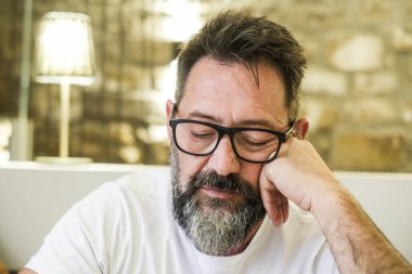 50 'li yaşlarda orta yaşlı bir adam evde tek başına oturuyor, gözlük takıyor ve derinden düşünüyor, yüzünde üzüntü, endişe ve duygusal stres belirtileri var.