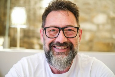 Sakallı ve gözlüklü bir adam evinde oturuyor, sıcak bir gülümsemeyle kameraya bakıyor, bulanık arka plan rahat ve güvenilir atmosferi arttırıyor.