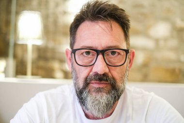 Ciddi görünümlü bir adam, evde duruyor, sert bir ifadeyle derinden kızgın görünüyor. Yüzü yoğun bir hayal kırıklığı ve stres gösteriyor, özel hayatında ve kariyerinde zorluklar var, genel olarak gergin bir atmosfer yaratıyor.