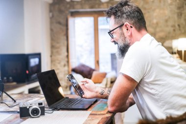 Orta yaşlı bir adam ofis masasında dizüstü bilgisayar ve akıllı telefon kullanarak oturuyor. Serbest fotoğrafçılık işinde uzaktan çalışıyor. Masasında bir kamera ve arka planda şömine var.
