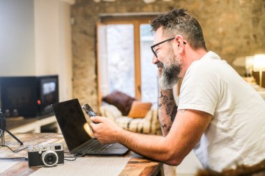 Çalışma odasında dizüstü bilgisayar ve akıllı telefon kullanarak serbest dijital çalışan, masadaki kanepe, şömine ve profesyonel fotoğraf ekipmanlarıyla modern oturma odasında uzaktan çalışan biri.