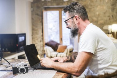 Orta yaşlı bir adam ofis masasında oturuyor, dizüstü bilgisayarı üzerinde çalışıyor, masa ve şömineli oturma odası arka planı, uzaktan çalışma konsepti, dijital meslek, modern çalışma alanı
