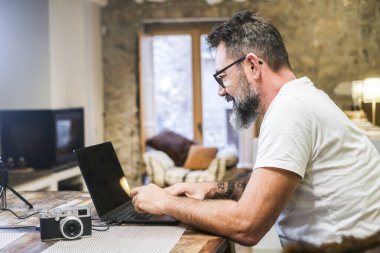 Günlük kıyafetli serbest çalışan ofis masasında dizüstü bilgisayarda çalışan, teknoloji, internet, uzaktan kumandalı iş, dijital iş, masada kamera, kanepe ve şömineli modern oturma odası