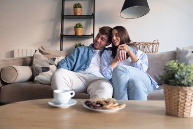 Genç bir çift, evlerinde rahat bir pazar öğleden sonrasında, kanepede oturuyor, konuşuyor, gülüyor, içki içiyor ve kurabiye yiyor, rahat bir atmosferi ve iletişim anları var.