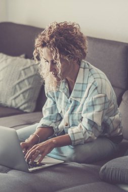 Evden uzakta çalışan bir kadın, dizüstü bilgisayarıyla bir koltukta oturuyor, internette geziniyor, online tarifleri araştırıyor, yeni yemekler okuyor, ve dijital iş aktivitelerine katılıyor.