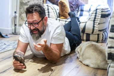 50 yaşında bir adam evinin zemininde uzanıyor, akıllı telefonunu sosyal medyada gezinmek için kullanıyor.