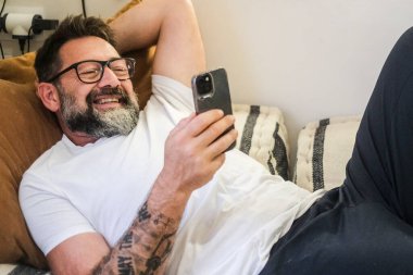 Yetişkin bir adam evdeki kanepede rahat bir şekilde oturup, internet içeriğine, sosyal medya kaynaklarına veya mesajlaşma uygulaması üzerinden sohbet ederken rahat bir ifadeyle akıllı telefonunu kullanıyor.