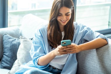 Genç kız evde kanepede otururken gülümsüyor, akıllı telefonu sosyal medya için kullanıyor, çevrimiçi sohbet ediyor, bilgi araştırıyor, dijital içeriği okuyor, hizmetlere abone oluyor ve internette geziniyor.