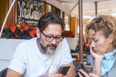 Her ikisi de 48 yaşında olan bir çift, duygusal uzaklıklarla ayrılmış bir restoranın açık hava yemek alanında otururken, karşılıklı akıllı telefonlarına odaklanıyorlar, etkileşim veya konuşmadan kaçınıyorlar..