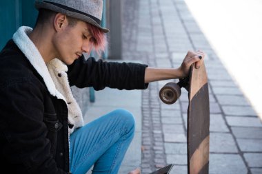 Genç bir erkek, şehir merdivenlerinde kaykay tutarken, gündelik bir kıyafetle, düşüncelere dalmış, şehir dışında kaybolmuş bir halde.