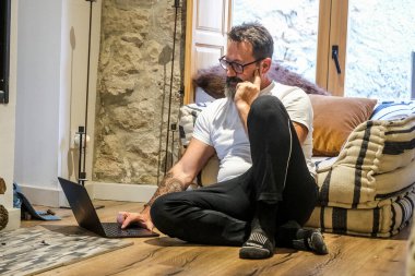 50 yaşında bir adam dağ evinin zemininde oturmuş, internette gezinip sosyal medyayı kontrol ederken huzur içinde dinleniyor ve derin derin düşünüyor.