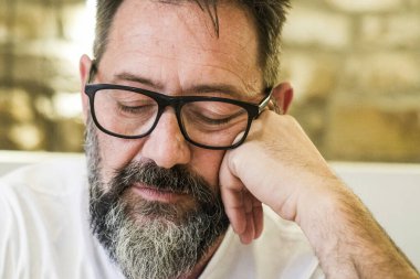 50 'li yaşlarda orta yaşlı bir adam evde tek başına oturuyor, gözlük takıyor ve derinden düşünüyor, yüzünde üzüntü, endişe ve duygusal stres belirtileri var.