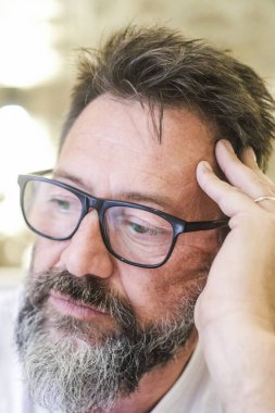 50 yaşlarında orta yaşlı bir adam, evde tek başına oturuyor, gözlük takıyor, yüzünde endişe ifadesi, derin düşünce ve zihinsel yorgunluk var.