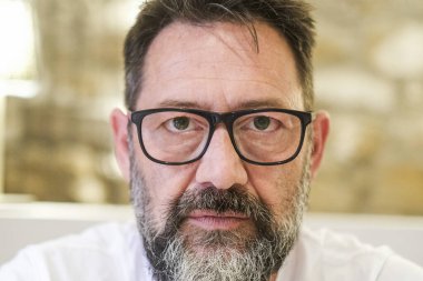 Ciddi görünümlü bir adam, evde duruyor, sert bir ifadeyle derinden kızgın görünüyor. Yüzü yoğun bir hayal kırıklığı ve stres gösteriyor, özel hayatında ve kariyerinde zorluklar var, genel olarak gergin bir atmosfer yaratıyor.