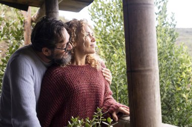 Mutlu bir yetişkin çift balkonda birlikte sakin bir anın tadını çıkarır, doğayla iç içe, birlikte parlak bir gelecek düşlerken birbirlerini kucaklarlar, kiralık dairelerinde huzurlu bir tatil yaşarlar.