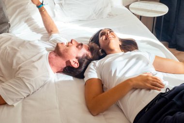 Yatağın üzerinde oturan bir çift, tamamen giyinmiş, gülüyor ve birlikte eğleniyorlar, neşeli gülümsemeler takınıyorlar ve rahat bir ortamda eğlenceli bir sohbet ediyorlar.