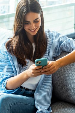 Gülümseyen genç bir milenyum kadını koltukta oturmuş, rahat, akıllı bir telefon tutuyor, sohbet etmek, internette gezinmek, online hizmetlere abone olmak, flört etmek gibi dijital aktivitelere katılıyor.