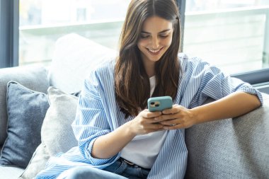 Genç kız evde kanepede otururken gülümsüyor, akıllı telefonu sosyal medya için kullanıyor, çevrimiçi sohbet ediyor, bilgi araştırıyor, dijital içeriği okuyor, hizmetlere abone oluyor ve internette geziniyor.