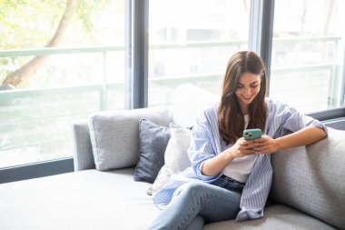 26 yaşında, uzun saçlı bir kadın oturma odasındaki gri bir kanepede rahatça oturuyor. Akıllı telefonuyla komik videolar izlerken gülümsüyor. Arkasında doğal ışık sağlayan büyük bir pencere var..