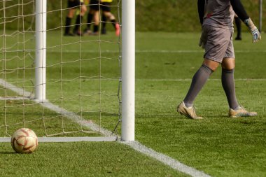 Sentetik çimlerle futbol sahası, sol altta bir top, kale direkleri, kale ağı ve arka planda üç hakem ile kaleci.