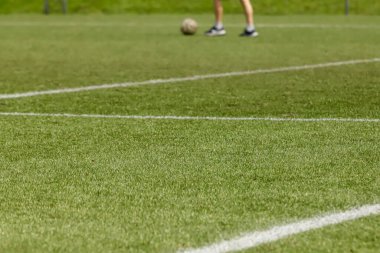Futbol sahası sentetik çimlerle, alt üçte ikisinde beyaz çizgiler ve üst üçüncüsünde topu olan bir sporcunun ayakları..