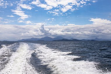 Bir sürat teknesinin motorunun izleri ön planda suda, arka planda bulutlar arasında mavi bir gökyüzü ve arka planda Serra do Mar.