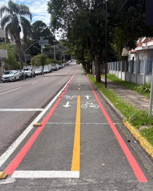 Asfaltta kırmızı, sarı ve beyaz bisiklet şekilli çizgileri olan bisikletçiler için özel bir alan. Sağ tarafta çimenler ve kaldırımlar var, sağda, park edilmiş arabalar ve her iki tarafta ağaçlar..