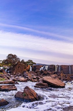 Arka planda güzel bir şelale olan kayalık bir sahilin dikey görüntüsü.