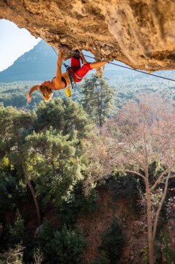 Kız kaya tırmanışçısı bir ormanın arka planına ve mavi dağlara tırmanır. Dağcı ekipman kullanarak zor bir yoldan geçiyor. Türkiye 'de kaya tırmanışı.