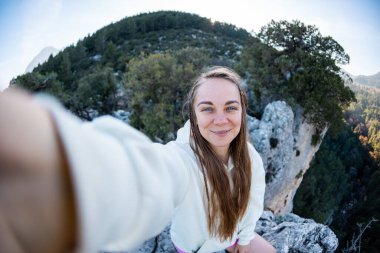 Young woman with blond hair taking a selfie portrait while hiking in the mountains - Happy hiker on top of a cliff smiling at camera - Travel and hobby concept