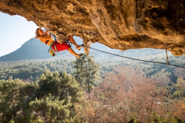 Kız kaya tırmanışçısı bir ormanın arka planına ve mavi dağlara tırmanır. Dağcı ekipman kullanarak zor bir yoldan geçiyor. Türkiye 'de kaya tırmanışı.