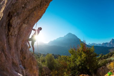 Atletik bir adam, sarkık bir kayaya halatla tırmanır, kurşun tırmanışı yapar. Dağ zemininde bir kaya tırmanıcısının silüeti. Açık hava sporları ve eğlence