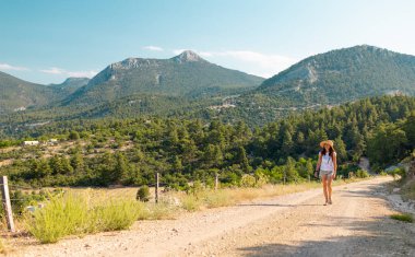 girl traveler with a backpack goes along a mountain path. a young girl in a hat with a backpack walks along the road in the forest against the backdrop of mountains and blue sky