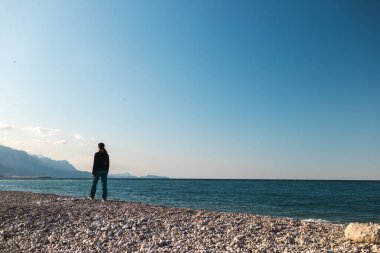 Bir kız sahilde tek başına yürüyor, bir kadın denize bakıyor, deniz ve dağların arkasında esmer bir kadın, deniz kıyısında dinleniyor.